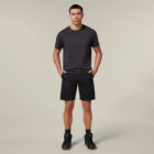 A person confidently stands against a neutral background in a dark T-shirt and shorts, wearing black sneakers. They hold an Emz A5 Notebook in one hand while the other rests casually in their pocket, striking a relaxed pose.