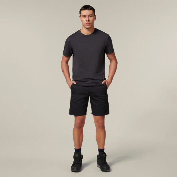 A person confidently stands against a neutral background in a dark T-shirt and shorts, wearing black sneakers. They hold an Emz A5 Notebook in one hand while the other rests casually in their pocket, striking a relaxed pose.