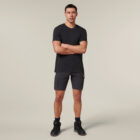 A person in a black T-shirt, gray cargo shorts, and black boots stands against a plain background, holding an Emz A5 Notebook with arms crossed. Their short, dark hair adds to the look.