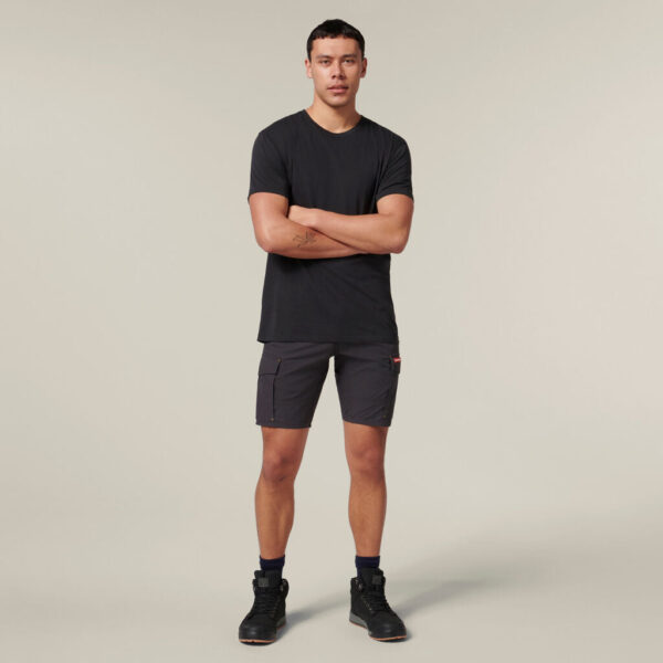 A person in a black T-shirt, gray cargo shorts, and black boots stands against a plain background, holding an Emz A5 Notebook with arms crossed. Their short, dark hair adds to the look.