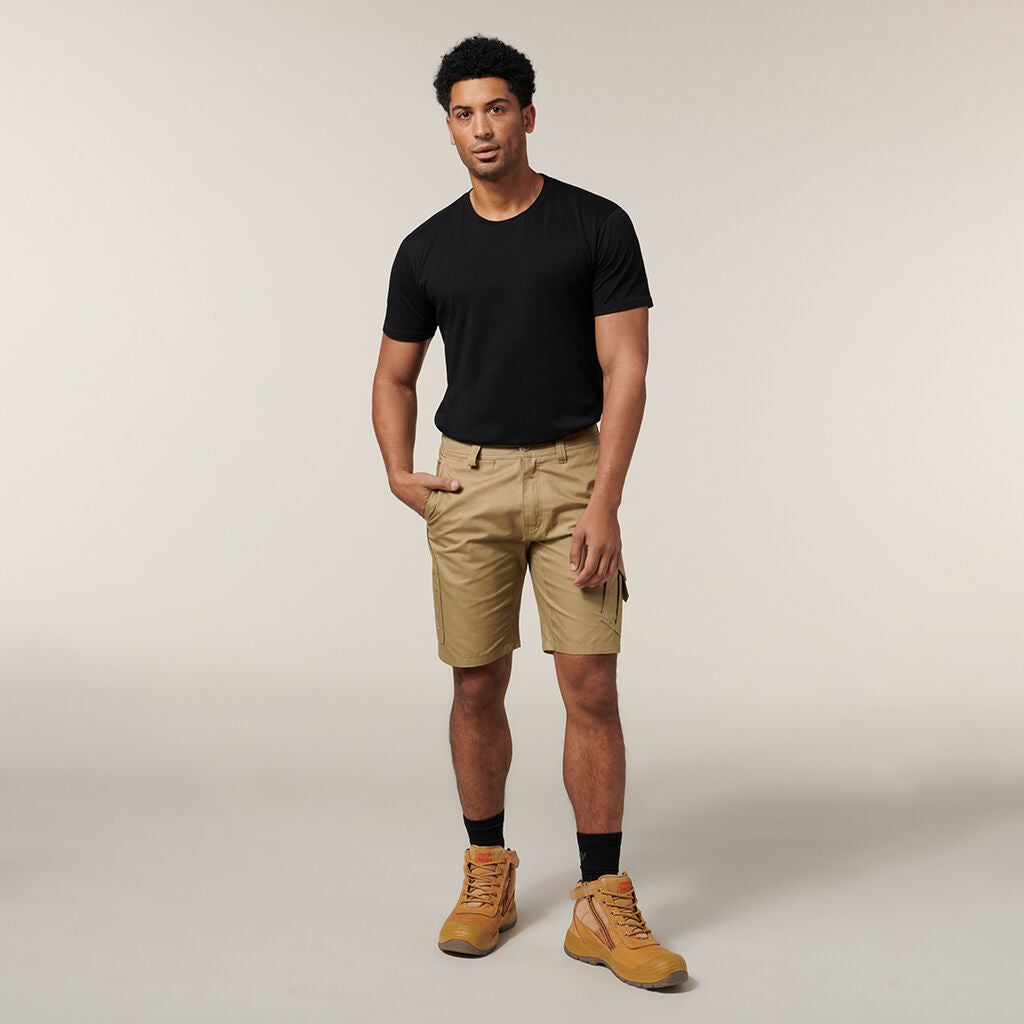 A man in a black t-shirt, beige cargo shorts, black socks, and tan boots stands casually with hands in pockets against a plain background. The Emz A5 Notebook beside him adds practicality to his relaxed style.