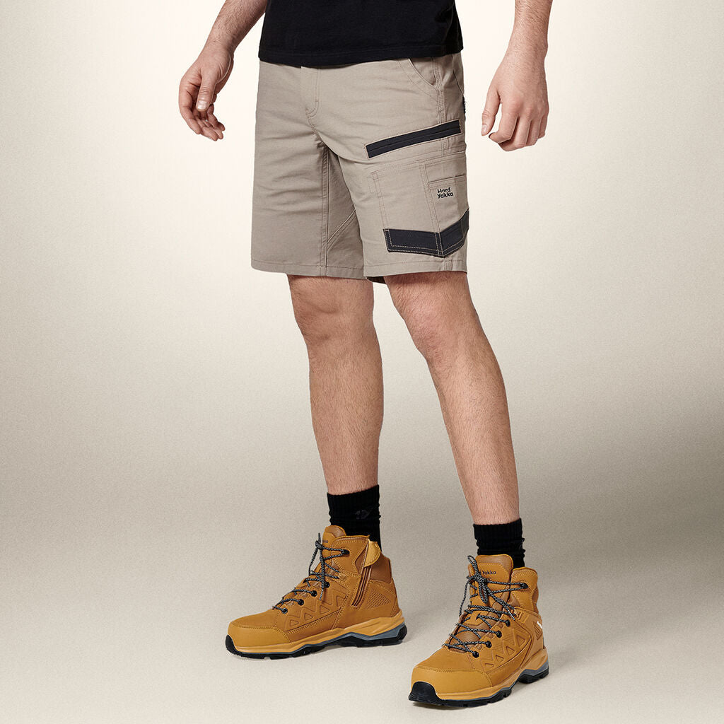 A person dons beige cargo shorts, a black t-shirt, black socks, and brown hiking boots. The shorts feature a left-side pocket with a black logo, ideal for storing an Emz A5 Notebook. The backdrop is a plain light color.