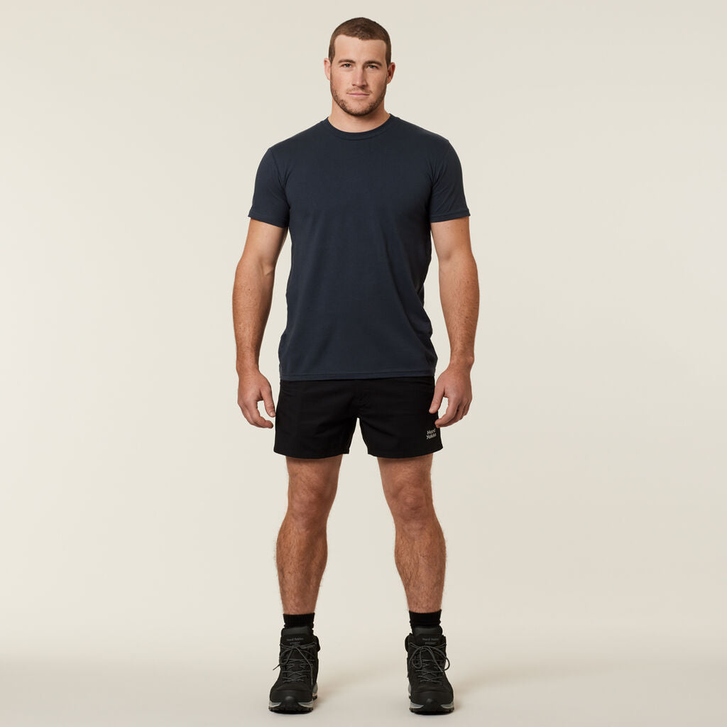Against a plain backdrop, a man stands facing forward in a dark t-shirt, black shorts, and sneakers with socks. He adds personality to his simple outfit by casually holding an Emz A5 Notebook.
