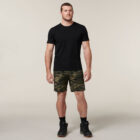 A man stands against a plain background wearing a black T-shirt, camo shorts, black socks, and shoes. Holding an Emz A5 Notebook under one arm, his neutral expression and relaxed stance exude quiet confidence.