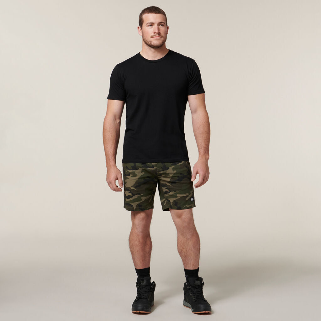 A man stands against a plain background wearing a black T-shirt, camo shorts, black socks, and shoes. Holding an Emz A5 Notebook under one arm, his neutral expression and relaxed stance exude quiet confidence.