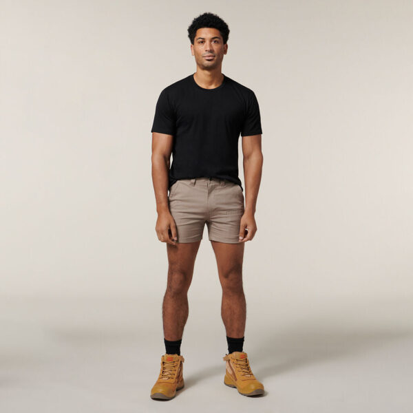 An individual in a black T-shirt, beige shorts, black socks, and tan boots stands against a plain background holding an Emz A5 Notebook. They have short dark hair, a neutral expression, and relaxed arms.
