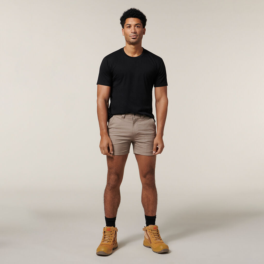 An individual in a black T-shirt, beige shorts, black socks, and tan boots stands against a plain background holding an Emz A5 Notebook. They have short dark hair, a neutral expression, and relaxed arms.