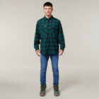 Against a neutral background, a person wearing a green and black plaid shirt, blue jeans, and green boots holds an Emz A5 Notebook. Their short dark hair completes their relaxed yet thoughtful appearance.