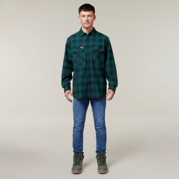 Against a neutral background, a person wearing a green and black plaid shirt, blue jeans, and green boots holds an Emz A5 Notebook. Their short dark hair completes their relaxed yet thoughtful appearance.