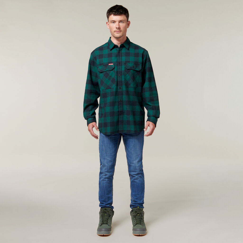 Against a neutral background, a person wearing a green and black plaid shirt, blue jeans, and green boots holds an Emz A5 Notebook. Their short dark hair completes their relaxed yet thoughtful appearance.