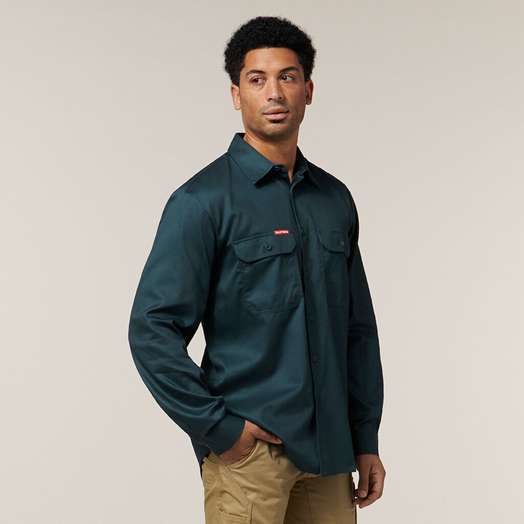 A person in a dark green button-up shirt with two chest pockets and tan pants stands against a plain background, holding an Emz A5 Notebook. They have short, curly hair and gaze slightly to the side.