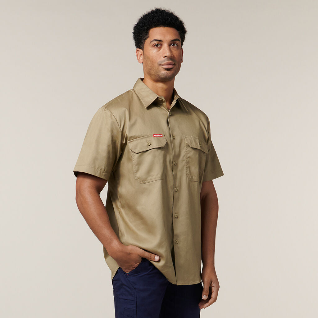 A person with short curly hair, wearing a short-sleeve tan button-up shirt and dark pants, stands against a plain background with one hand in their pocket, casually holding an Emz A5 Notebook.