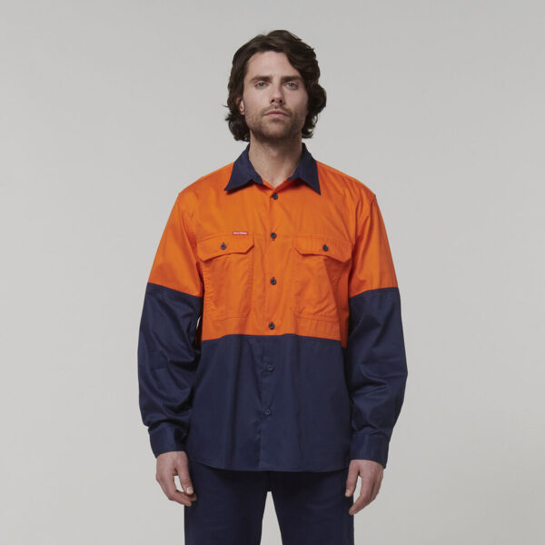 A man with medium-length dark hair wears a long-sleeve work shirt, orange on top and navy blue below, along with matching pants. He holds an Emz A5 Notebook in his hand and stands against a neutral background, looking directly at the camera.