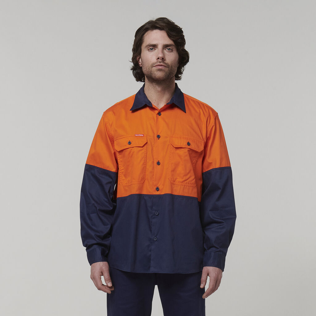 A man with medium-length dark hair wears a long-sleeve work shirt, orange on top and navy blue below, along with matching pants. He holds an Emz A5 Notebook in his hand and stands against a neutral background, looking directly at the camera.