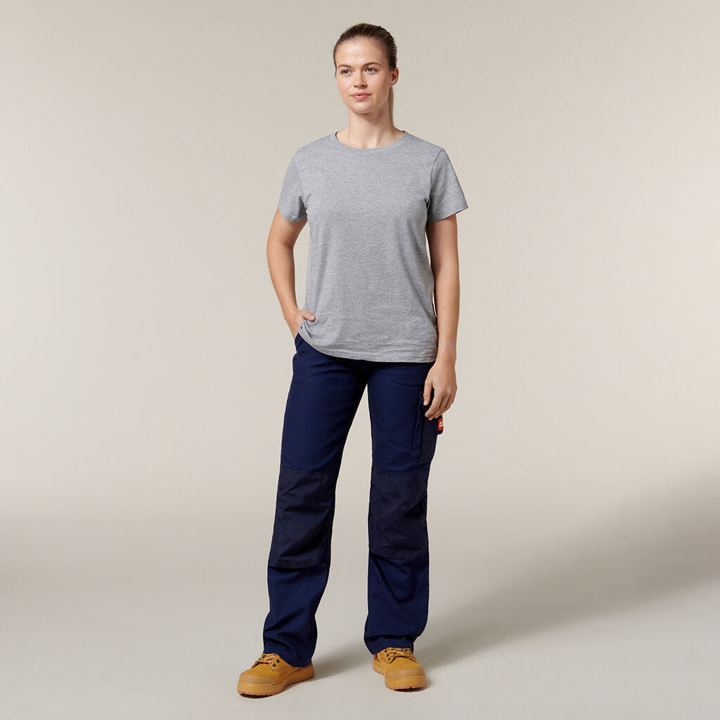 A person in a gray t-shirt and dark blue cargo pants stands against a plain background, casually holding an Emz A5 Notebook. They wear tan work boots and have short hair, looking ahead with a calm expression.
