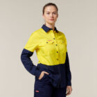In an Emz signature high-visibility uniform featuring a bright yellow top with dark blue sleeves and pants, a person confidently stands with one hand in their pocket against a plain, off-white background.