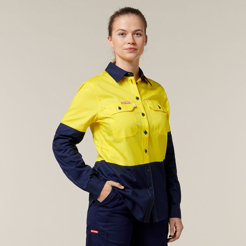 In an Emz signature high-visibility uniform featuring a bright yellow top with dark blue sleeves and pants, a person confidently stands with one hand in their pocket against a plain, off-white background.