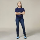 A person stands confidently with arms crossed in a navy blue t-shirt, blue jeans, and light blue high-top sneakers. The Emz A5 Notebook beside them adds a creative touch to the plain neutral background.