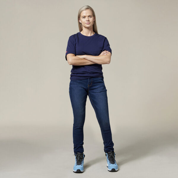 A person stands confidently with arms crossed in a navy blue t-shirt, blue jeans, and light blue high-top sneakers. The Emz A5 Notebook beside them adds a creative touch to the plain neutral background.