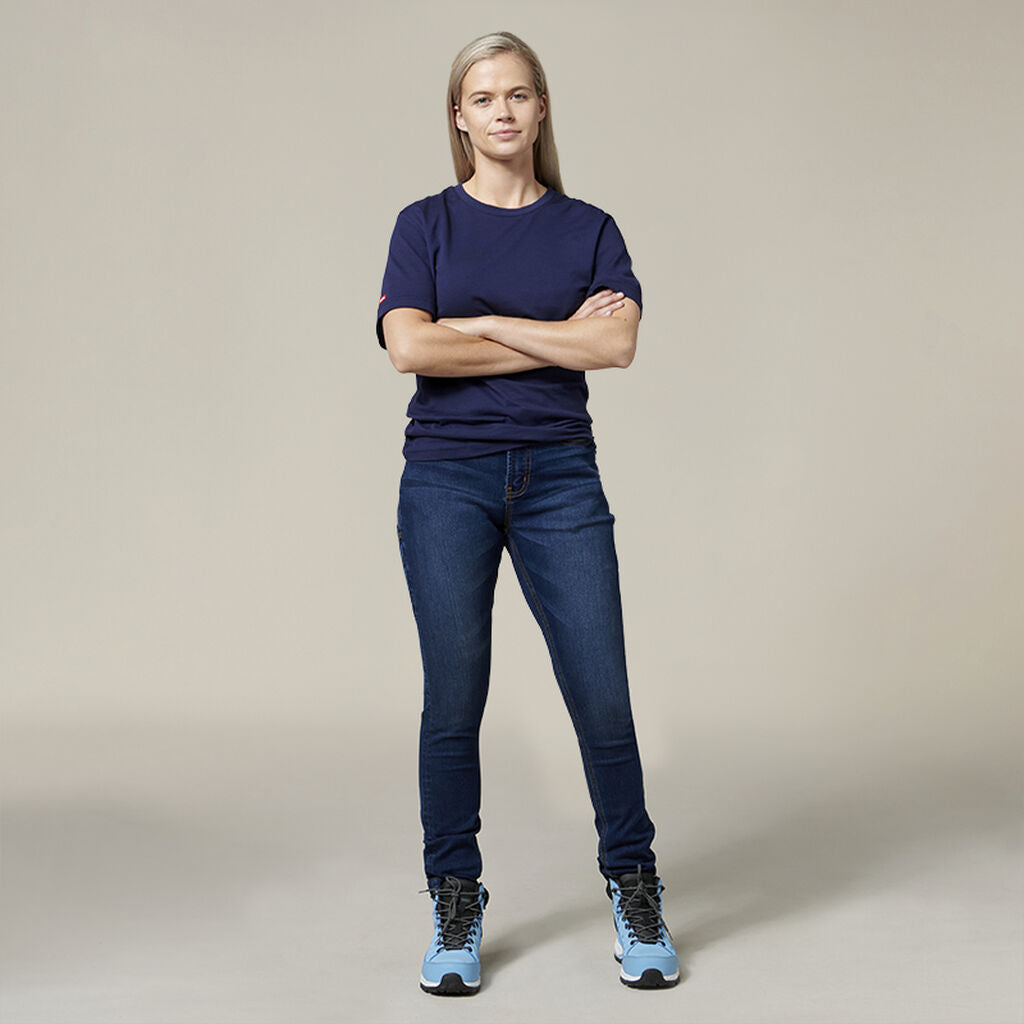 A person stands confidently with arms crossed in a navy blue t-shirt, blue jeans, and light blue high-top sneakers. The Emz A5 Notebook beside them adds a creative touch to the plain neutral background.
