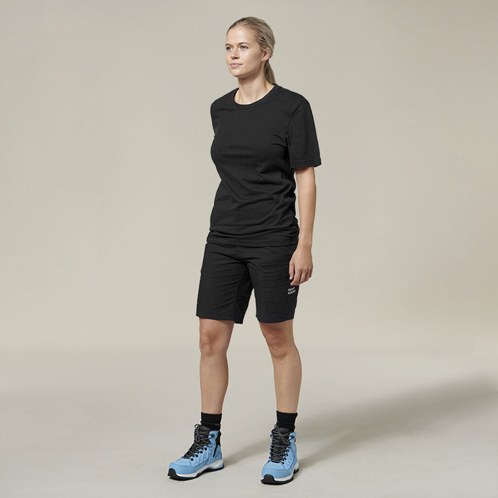 A person dressed in a black T-shirt, black shorts, and light blue hiking boots stands against a plain backdrop, looking to the side with an Emz expression suggesting deep thought, as if contemplating entries in an Emz A5 Notebook.