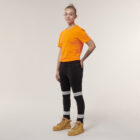 A person with short hair in an orange T-shirt and black jeans stands against a plain background. Wearing tan boots, they display a tattoo on one arm and hold an Emz A5 Notebook that complements their style.