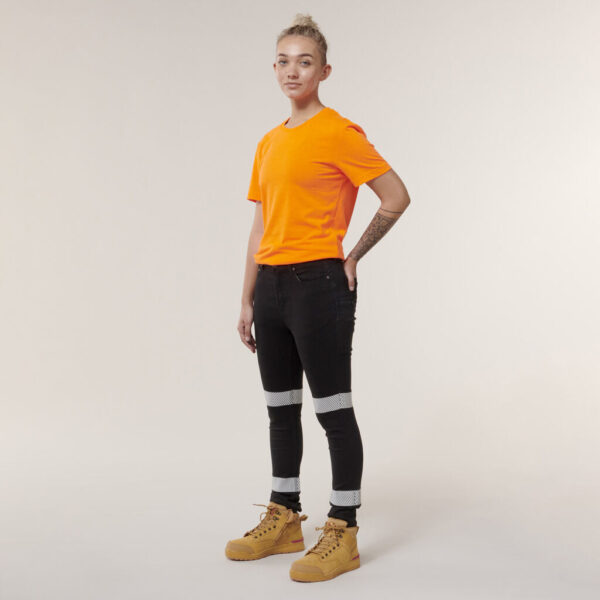 A person with short hair in an orange T-shirt and black jeans stands against a plain background. Wearing tan boots, they display a tattoo on one arm and hold an Emz A5 Notebook that complements their style.