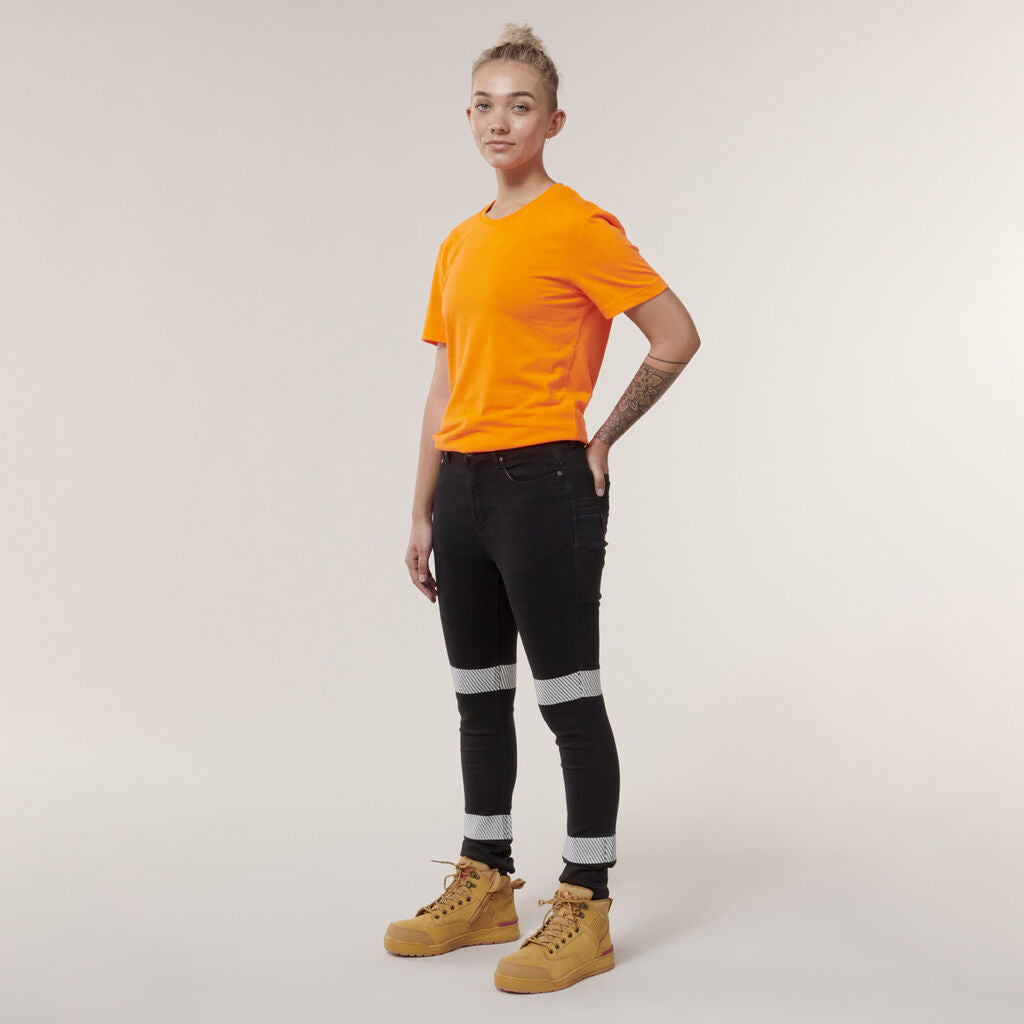 A person with short hair in an orange T-shirt and black jeans stands against a plain background. Wearing tan boots, they display a tattoo on one arm and hold an Emz A5 Notebook that complements their style.