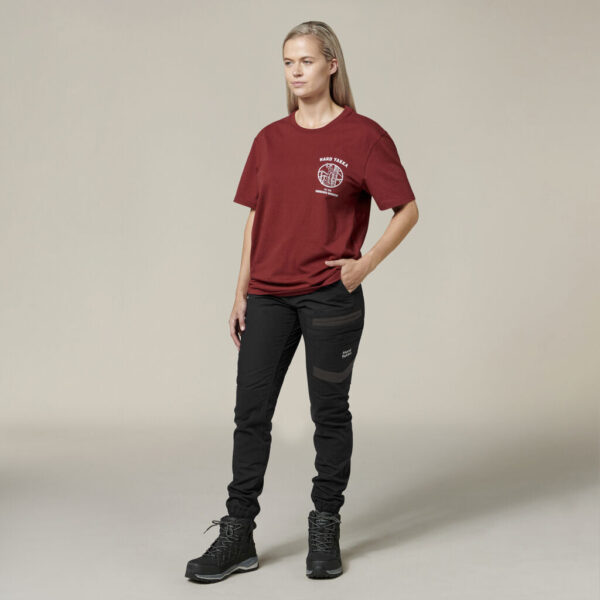 A person stands neutrally against a plain backdrop, wearing a maroon T-shirt adorned with Emzs logo. They pair it with black pants that have pocket detailing and dark hiking boots, casually holding an Emz A5 Notebook in one hand.