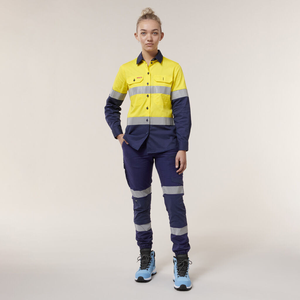Decked in a vibrant yellow and dark blue work uniform adorned with reflective stripes, the person confidently poses against a plain background. They hold an Emz A5 Notebook in hand and sport blue boots.