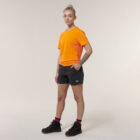 A confident person stands with one hand in their pocket, sporting an orange T-shirt, black shorts, and boots. A tattoo adorns their arm as they hold an Emz A5 Notebook against a light background, capturing both style and creativity.
