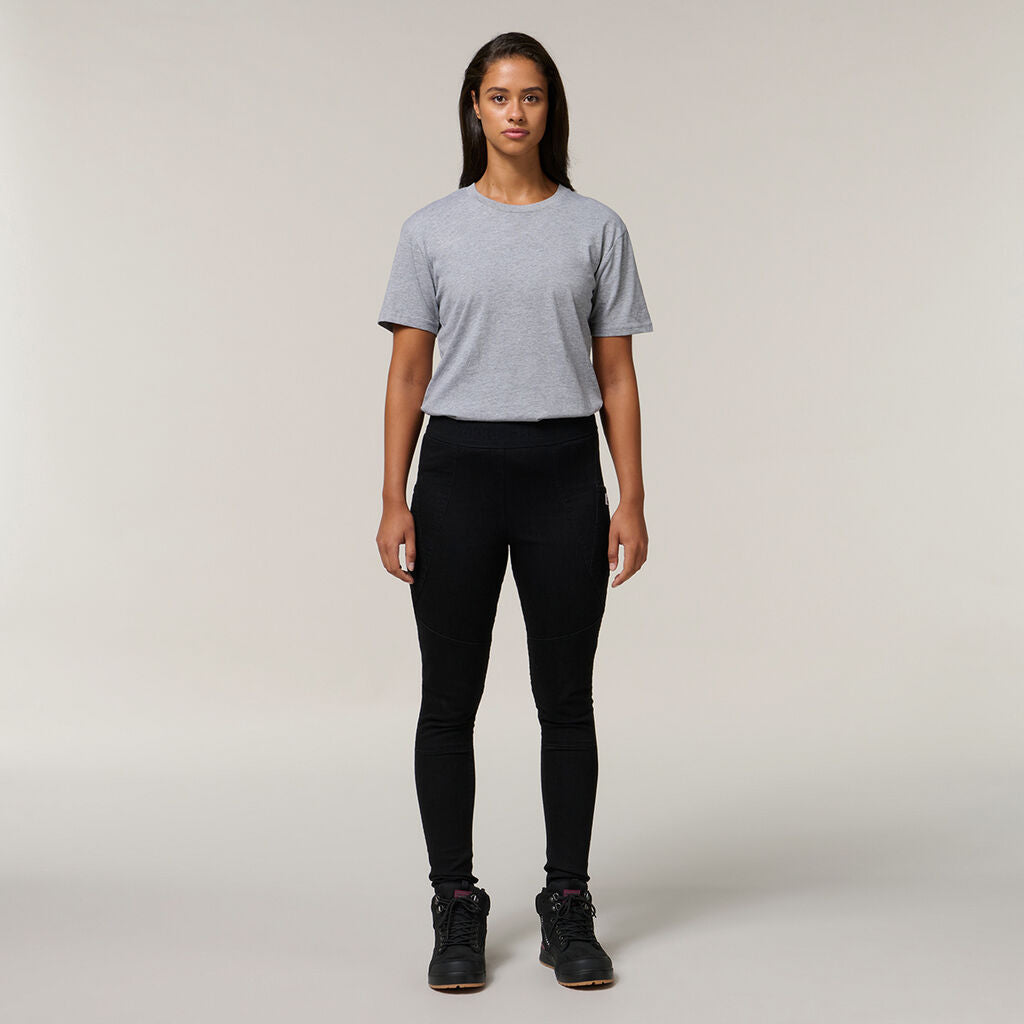 A person with long dark hair and a neutral expression stands against a plain background, holding an Emz A5 Notebook in one hand. Dressed in a gray t-shirt, black pants, and black shoes, their other arm rests by their side.