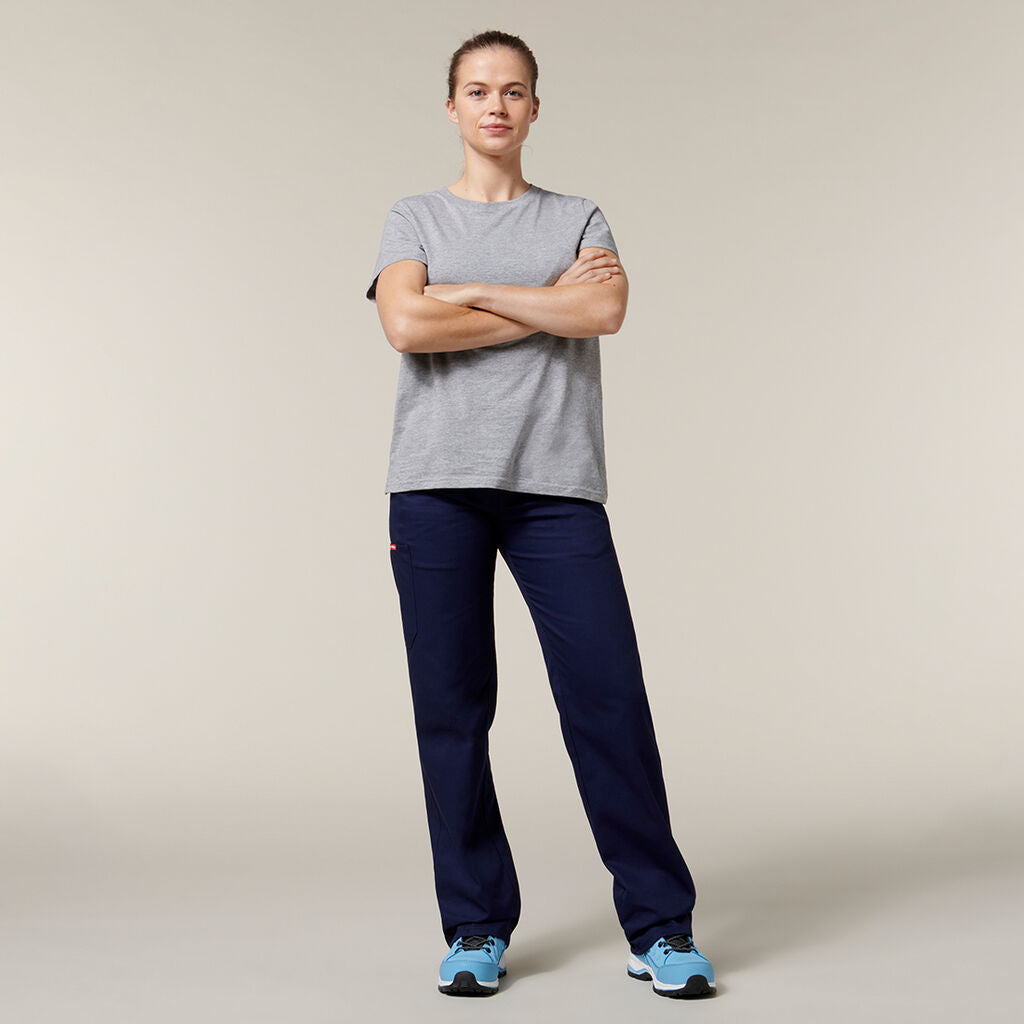 A confident individual stands with arms crossed, wearing a gray t-shirt, navy blue pants, and blue sneakers, holding an Emz A5 Notebook against a plain neutral background.