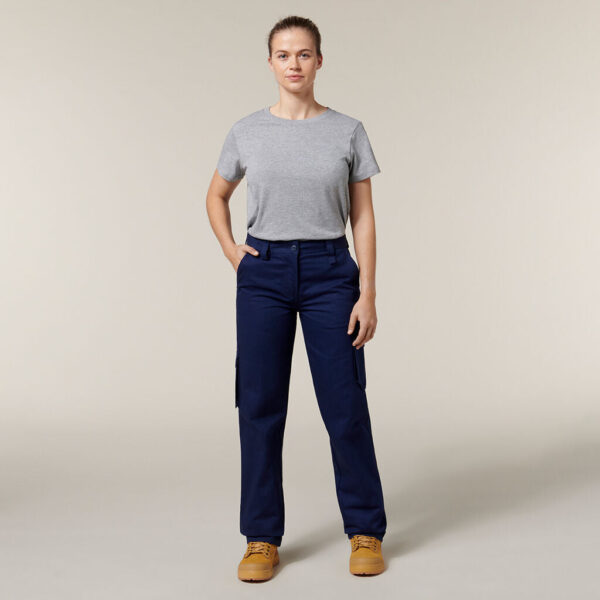 A confident person stands with hands in pockets, wearing a gray T-shirt and blue cargo pants. Clutching an Emz A5 Notebook under one arm, they have light brown hair tied back and sport tan work boots, all against a simple light beige backdrop.