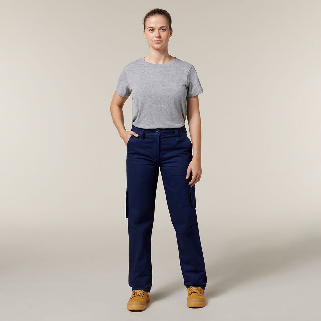 A confident person stands with hands in pockets, wearing a gray T-shirt and blue cargo pants. Clutching an Emz A5 Notebook under one arm, they have light brown hair tied back and sport tan work boots, all against a simple light beige backdrop.