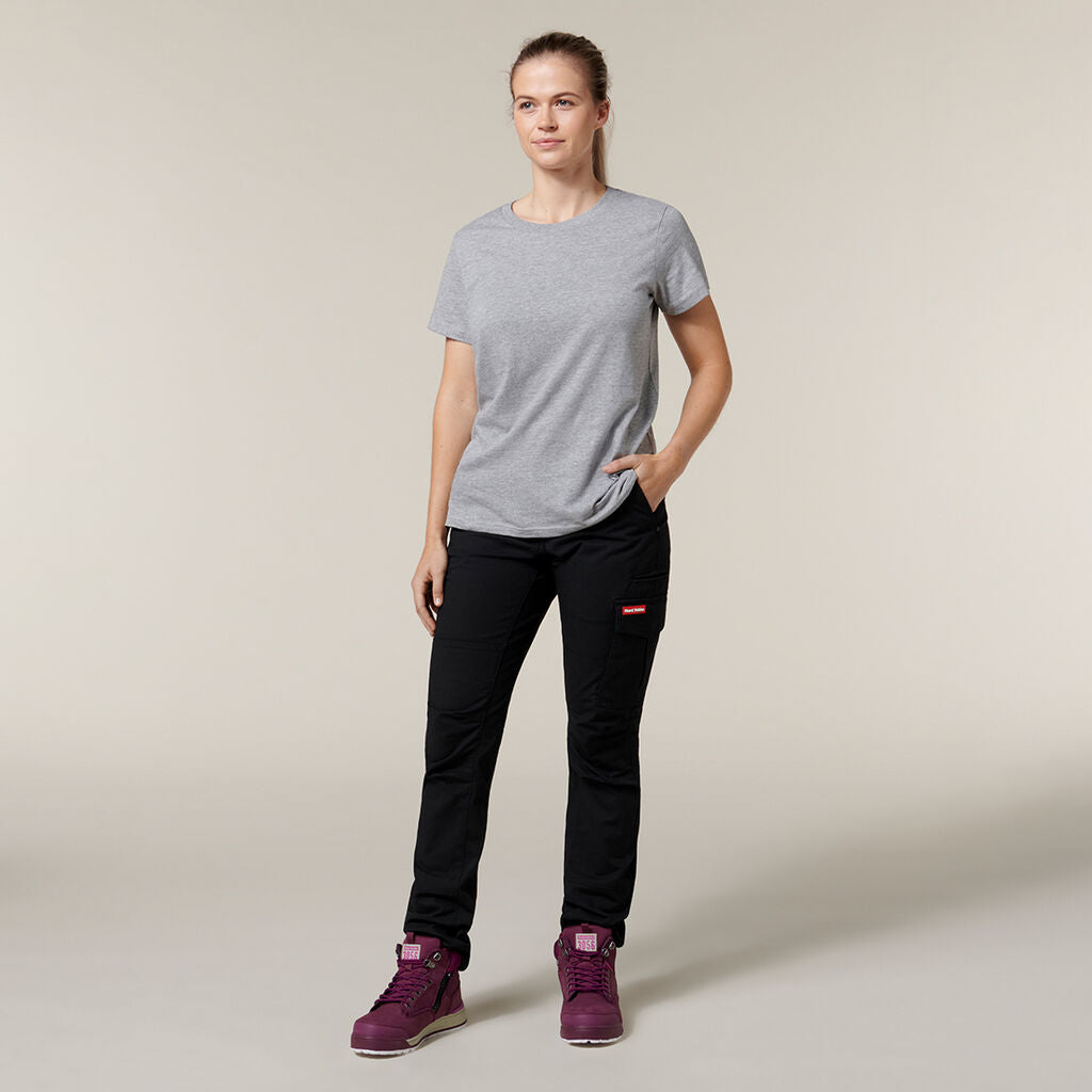 In front of a plain backdrop, someone sports a gray t-shirt, black pants, and purple high-tops. With one hand resting in their pocket and the other under their arm holding an Emz A5 Notebook—perfect for jotting down spontaneous ideas—they stand with a neutral expression.