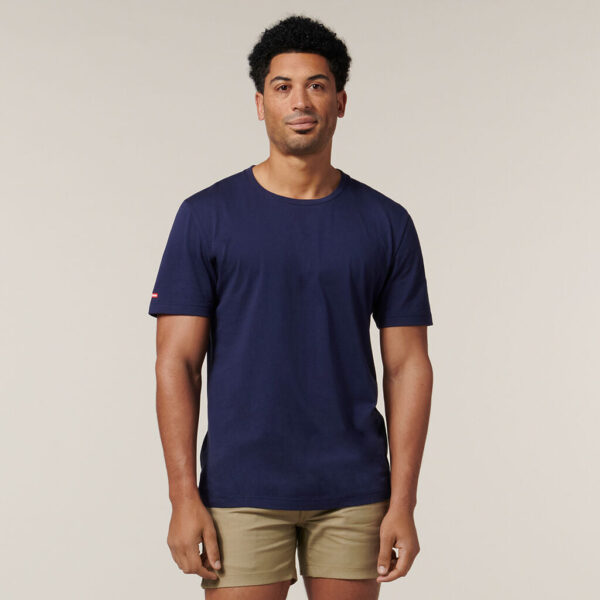 A person with short curly hair, in a navy t-shirt and khaki shorts, stands against a beige background holding an Emz A5 Notebook and faces the camera neutrally.