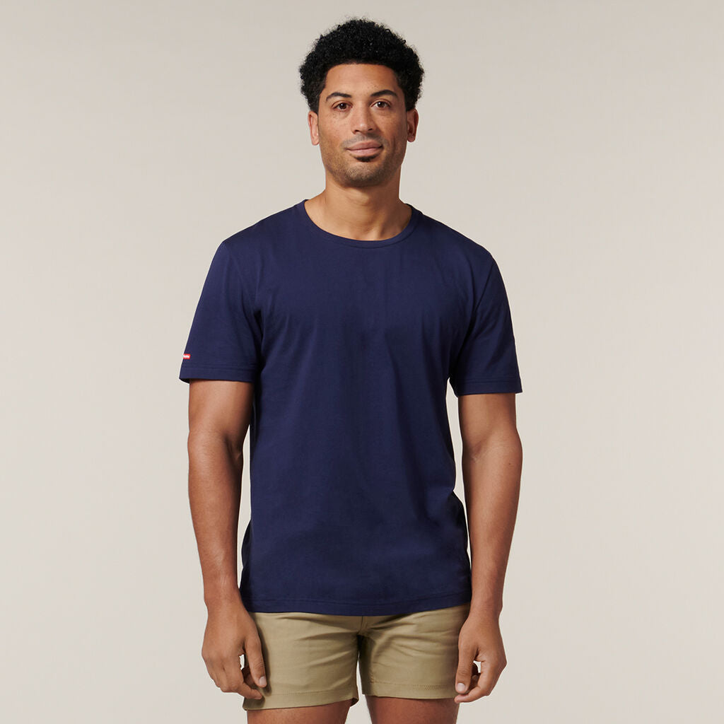 A person with short curly hair, in a navy t-shirt and khaki shorts, stands against a beige background holding an Emz A5 Notebook and faces the camera neutrally.