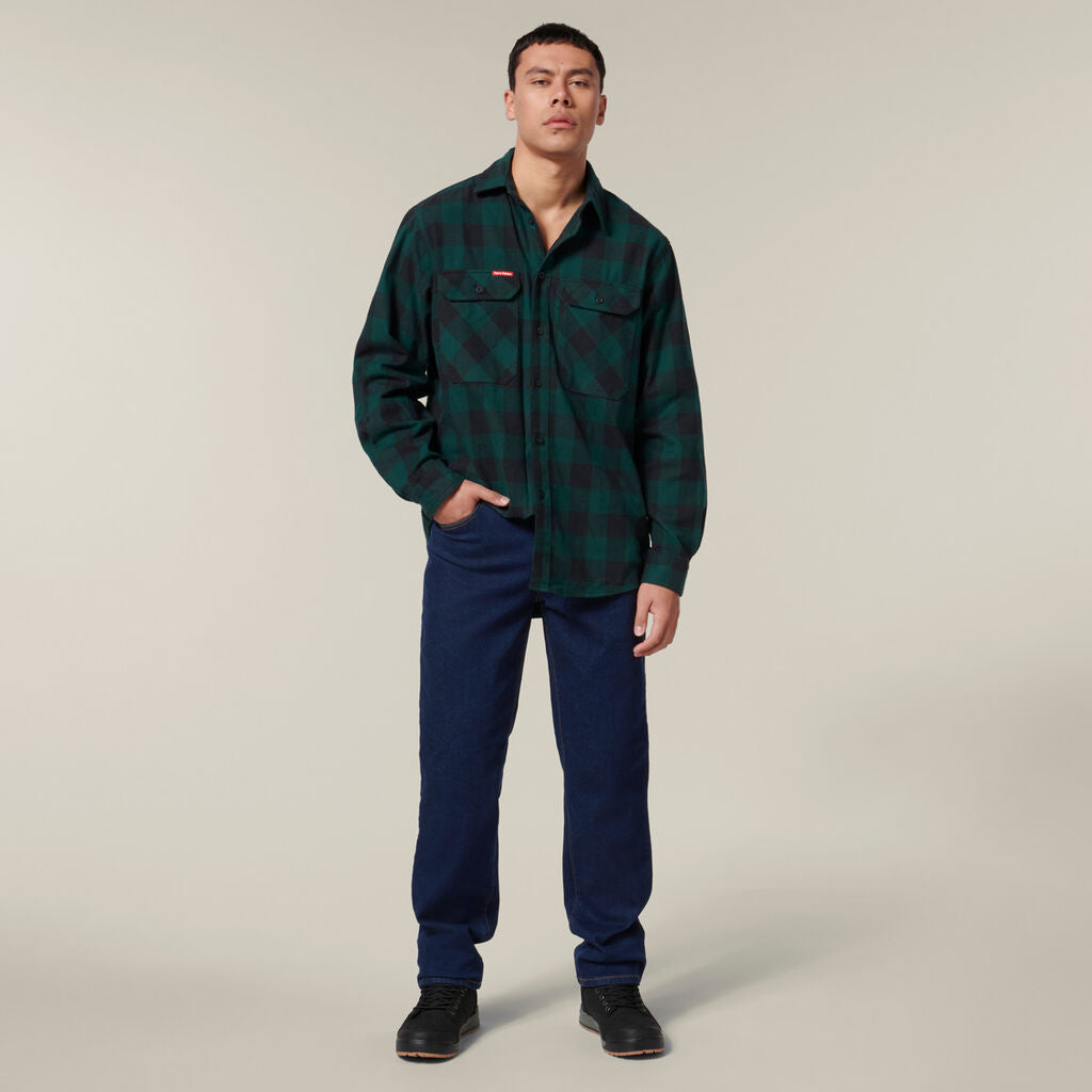 A person with short hair stands against a plain, light background, wearing a green and black checkered flannel shirt and dark blue pants. They are holding an Emz A5 Notebook while looking at the camera.