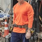 Man in orange shirt and tool belt stands at a construction site with equipment and debris.