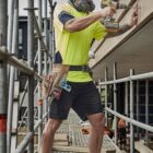 Construction worker in safety gear drills into wood on a scaffold at a building site.