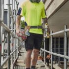 Construction worker in safety gear walking on scaffolding, holding tools at a building site.