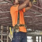 A construction worker in safety gear uses a power drill on a ceiling framework.