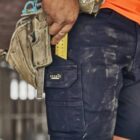 Person in work shorts with a tool belt and dusty hands holding a measuring tape.