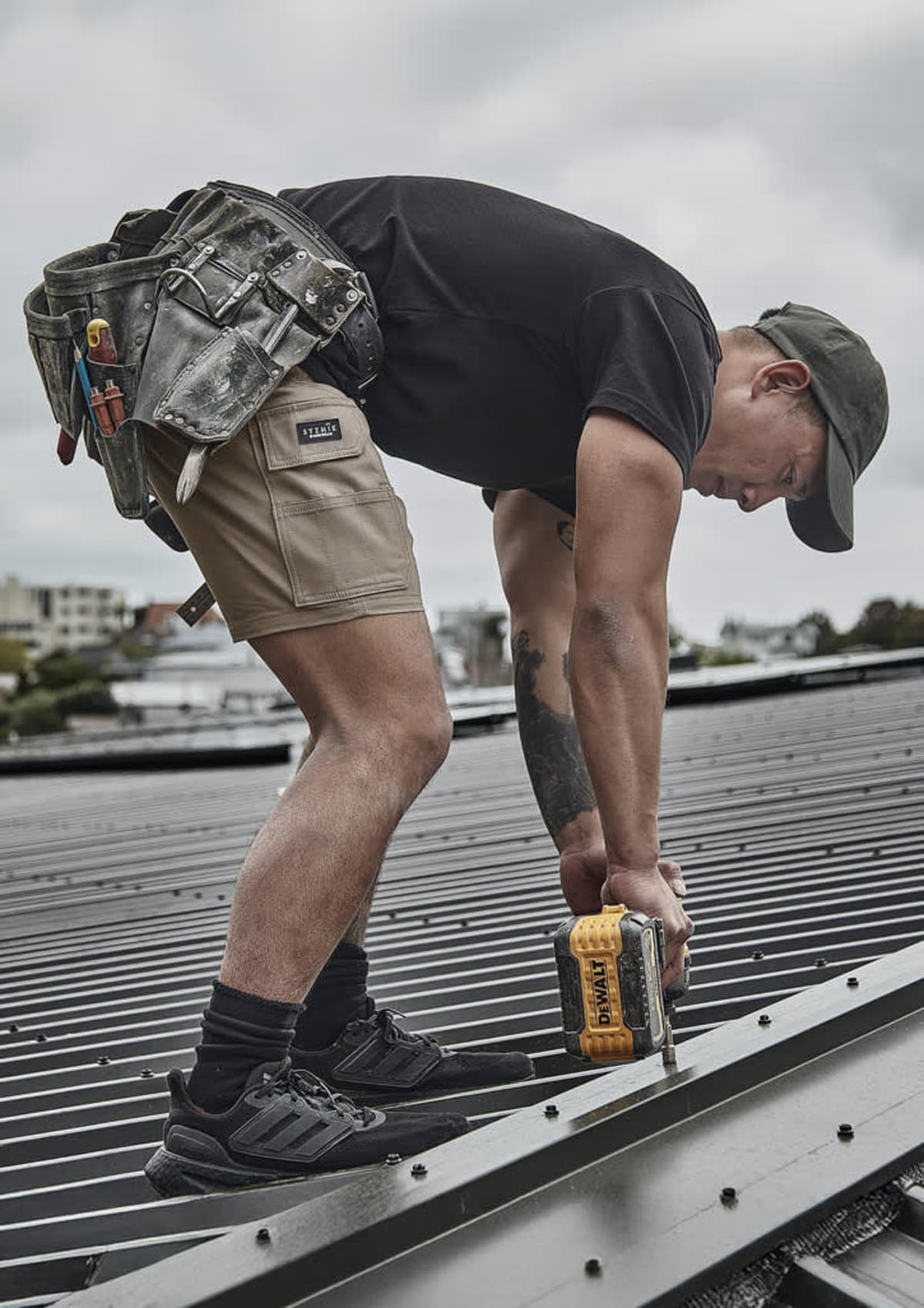 A construction worker drills into a metal roof while wearing shorts and a tool belt.