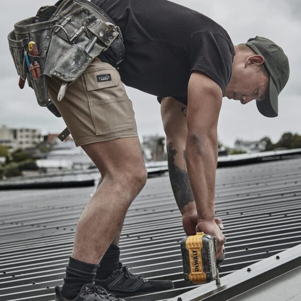 Man using a drill on a metal roof, wearing work shorts, tool belt, and a cap.