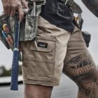 Person wearing tool belt and shorts, holding hammer, with tattooed leg visible.