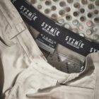 Beige SYZMIK workwear pants with visible waistband and brand labels on a metal surface.