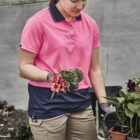 Woman in a pink polo shirt holding a flower and gardening outdoors by a table with plants.