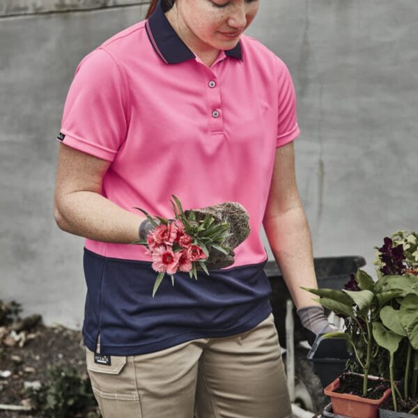 Woman in a pink polo shirt holding a flower and gardening outdoors by a table with plants.